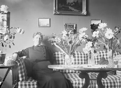 Dam i soffa med blomvaser på bordet. Göta Tisell; Ägare till huset Solliden.
