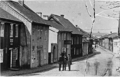 Storgatan väster. Längst bort i bakgrunden syns Väderkvarnen som fanns vid Åbro. Den flyttades till Öland, Sandvik och var färdig där 1885. Verkbyggmästare Grönkvist stod för nedmonteringen vilket han har beskrivit i sin dagbok som nu finns hos Fabrikör Karl Malmberg, Vimmerby. I Grankvistgården fanns då stadens siste tenngjutare Zilof, hans affärsskylt syns på bilden i form av 5 tenntallrikar, se även ett annat foto som togs samtidigt där man ser Storgatan från andra hållet, vklba3469. Lägg märke till att bilden är exponerad två gånger. De tre männen har rört på sig vid första exponeringen, då har fotografen satt på kameralocket och ordnat upp dem med anmaning att stå stilla, se även personen nere på Storgatan som har rört sig i sidled under tagningen.   T.E.
