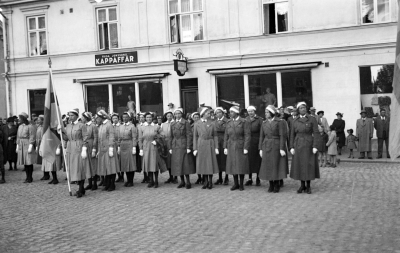 Flaggdagen,  Vimmerby Lottakår fanbärare Vivi Andersson, bakom fanbäraren fru "Putte Karlberg" 5. från vänster Karin Jönsson, Werngren, 1. från höger Nina Hansson, 2. från höger Maj Severin, 3. från höger Gunborg Lind 5. från höger Tina Dalin. 6. från höger Dagmar Gyllenkrok . Fastigheten vid Stora Torget  numera Sandströms. 1. Frida Jonssons  2. Sköldborgs damfrisering.



Dok.080205/RS