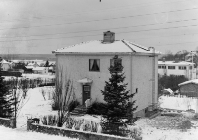 Östra Skolgatan 10,  hörnet Olle Persgatan/Östra Skolgatan som det såg ut innan nuvarande hyreshus byggdes. Nu återstår stenmuren, lite av syrenhäcken och den pampiga björken i hörnet. Greta Vogel ägde huset till 1975. Kortet bör vara taget från Sven "Esse" och Vera Ekbergs Balkong. De bodde mittemot på Östra Skolgatan. Till vänster syns gärdet vid Ringvägen, ovanför Källängsparken mittemot  dåvarande Centralbryggeriet. Det vita funkishuset som syns bakom Ö Skolgatan 10, till höger, ägdes av fru (Agda?) Ansgarius-Svensson. Den låga byggnaden till höger är Sandströms garage. Gissar att kortet är taget någon gång sent 40-tal eller tidigt 50-tal.    B.V.