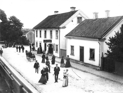Mannen i uniform är extrapolisen, rådhusvaktmästaren Axel Sköldborg. Storgatan Sekelskiftet 1900. Damen med ett barn på armen skall vara Fru L Sköldborg med dotter Ebba. Stora huset i mitten är Axel Eklunds affär. Lilla huset till höger är vaktmästarebostad för tingshusvaktmästare Sköldborg.   T.E.