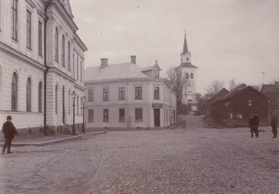 Bilden på Sevedegatan mot kyrkan är troligen tagen omkring 1890. I fastigheten i mitten av bilden drev E.M. Bauman klädaffär. Sonen Schamyl Bauman kom att bli en välkänd filmregissör. (Bildid. 18793)
