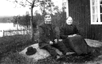 Carl Vilhelm Andersson, född 6/8-1847, död 1928, och hans hustru, Augusta Andersson, död 1920. Sjön Snesaren, Stugan kallas Björkhult och är stadsägan 4 i Vimmerby. Fastigheten äges numera fr.m. 1943 av Vivi Andersson Furugatan 5 B. Vimmerby. som är dotterdotter till Carl Vilhelm och Augusta Andersson. Fotografiet är troligen taget av Sigrid Tisell 1915 hon är syster till Anna och Göta Tisell, Vimmerby.    