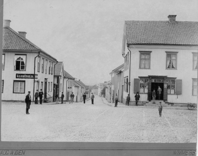 Storgatan v. om Torget. T.E.

Detta kort har jag fått vid bouppteckning den 12 / 3 1936 efter Ma ?  Sta ?
Kortet är från 1880 - talet 
Mannen till vänster med förkläde Karl Fransson, 8. från vänster Filip Dahl. Albert Karlsson son till nattvakt Karl Larsson, med rygg mot vägg under lucka, han körde ? ? med en enbetsoxe hos Fagerholm o Hedström. Vet intet annat namn än Pål, bodde i Potaskestugan. På nedre trappsteget ett biträde hos Barkman vid namn Arvidsson ?   ? från Mariannelund. Överst biträdet Nilsson han ? Band ? hade släktingar i Boda Frödinge, for sedan till Amerika ? har ?  ?  ? över Magkrampen. Pilten ut på torget heter Axl Dal ? han är idag hovmästare i Stockholm.
den 11 / 11 1949 Axel Almén.

Till vänster Kalmar Enskilda Bank och till höger Barkmans diverseaffär
