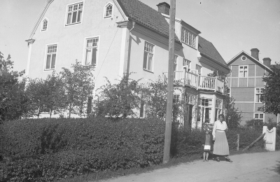 Drottninggatan 29 huset till vänster, Anna och Göta  Thysell ägde huset på 1930-talet. De hade inackordering för samskoleelever från anna ort. Barnet kan möjligen vara Gullan Johansson, adoptivbarn hos fröken Anna Johansson, lärare, i vackra röda huset, Drottninggatan 39. Damen är Anna Thysell. huset till höger Drottninggatan 31 är före detta Ernst Gustafssons speceriaffär.             