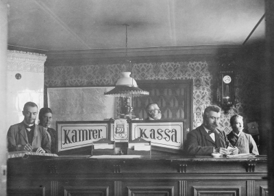  Barkmanska gården vid torget. I Barkmanska gården startades år 1910. Stockholms Köpmansbank upphörde 1913.  Skånska Handelsbanken 1919 övertogs 1949 av Skandinaviska, som nu heter Sveriges Kreditbank. Diskontobanken övertog Vimmerby Folkbank 1906, Startades 1899, Göteborgs bank övertog sedan Diskontobanken 1917 Göteborgs bank var i Sporres hus, när detta hus revs var banken placerad i motsvarande Systembolagets hus under byggnadstiden. Från vänster. 1 Kamrer ......?, 2 Elna Kjellström, 3 Vilhelm Westrin Direktör, 4 Valfrid Hägner Kassör, 5 Gustaf Jonsson Vaktmästare. Bilden tagen omkring 1904 i Barkmanska huset vid torget. 