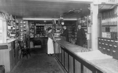 Fr.v. 1 Anna "Vale" Johansson Vimmerby, 2 John Svensson, Horn, handelbit, 3 Reinhold Berggren Handlande. 
Övrig dokumentation: Hökarboden i  Vimmerby drevs 1906 av Reinhold Berggren, en herre med psykologisk blick, tjuvpojken i ögat och hökeriet i själen. Det är han som står här närmast med sina medhjälpare Svante och Anna i bakgrunden, rapporterar Erik Lindström, 73 år, född i Vimmerby och bosatt i Stockholm. Själv samlade han som barn in gamla ben samt någon literbutelj, sålde dem hos lumphandlaren Rudman och köpte en strut karameller hos Berggren. "Med ABC-boken i ena fickan och literbuteljen i den andra tågade man till skolan glad och nöjd", meddelar hr Lindström. 