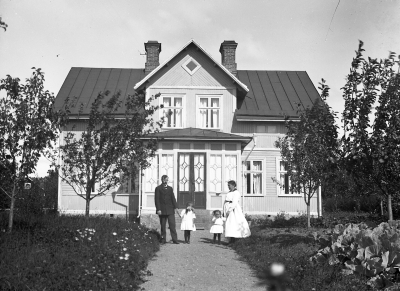 "Möbelsnickare Hjalmar Broo och hans familj framför fastigheten Milano på Loarpsgatan 8 i Vimmerby. Huset byggdes av änkefru Emilia Katarina Larsson f. Pettersson och hennes son Karl Julius Larsson 1909. Personerna fr.v är Hjalmar, döttrarna Gulli och Gertrud och hustrun Edit Karlsson"
