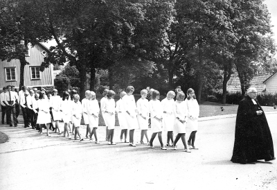 Sommarkonfirmander på väg från församlingshemmet till kyrkan.
Kyrkoherde Torsten Engström, nr 10 från höger, med långt ljust hår är Agneta Ståhl. Söndagen den 11 juli 1965. 